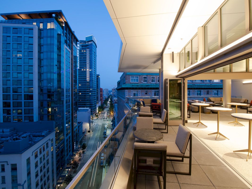 A modern cityscape at dusk with illuminated buildings, viewed from a balcony with tables and chairs, providing a stylish and inviting atmosphere.