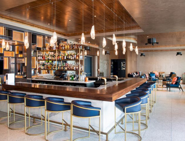This image shows a modern bar with wooden and metallic decor, featuring a well-stocked counter, stylish chairs, and ambient lighting fixtures.