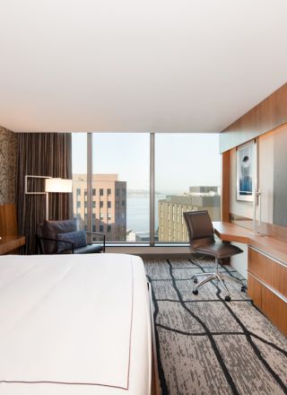 A modern hotel room with a large bed, desk, chair, TV, and a window offering a city view. The room features contemporary decor.