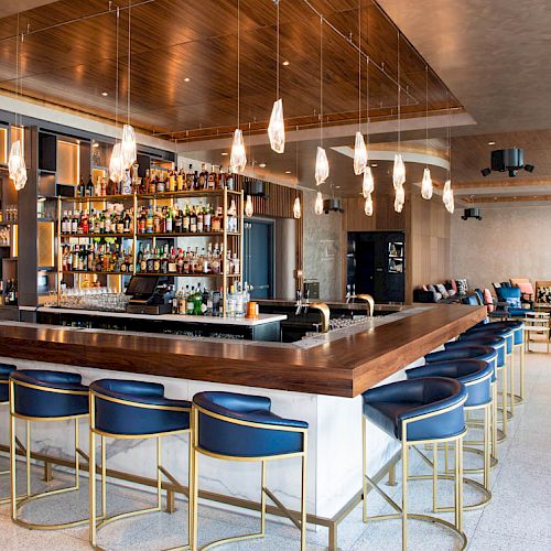 A modern bar with a sleek, wooden countertop, blue cushioned bar stools, and a well-stocked liquor shelf. Pendant lights hang from the ceiling.