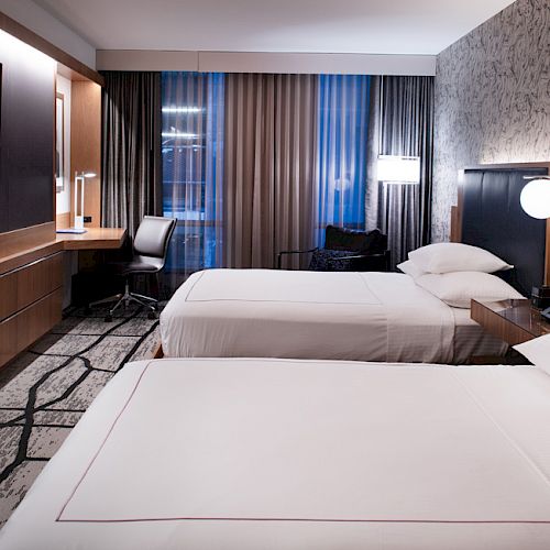 A modern hotel room with two beds, a wall-mounted TV, desk with a chair, bedside lamps, and a patterned wall, creating a comfortable ambiance.