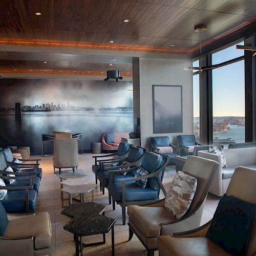 A modern, spacious lounge with armchairs, large windows offering waterfront views, and ambient lighting creating a cozy atmosphere.