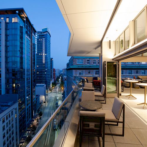 A modern cityscape at dusk with tall buildings and a lit-up balcony featuring seating and tables overlooking the streets below.