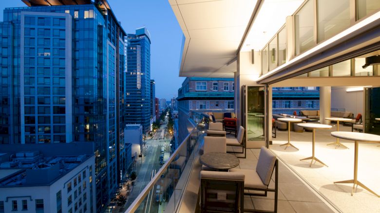 A modern cityscape at dusk, featuring tall buildings and a spacious balcony with seating and tables, overlooking a city street illuminated by lights.