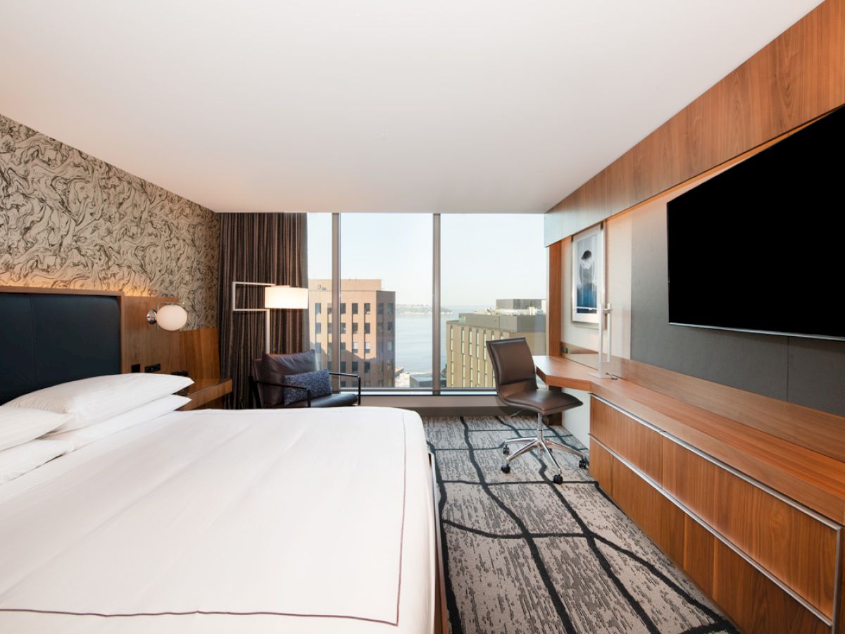 A modern hotel room with a large bed, city view through windows, a desk with a chair, mounted TV, and contemporary furnishings.