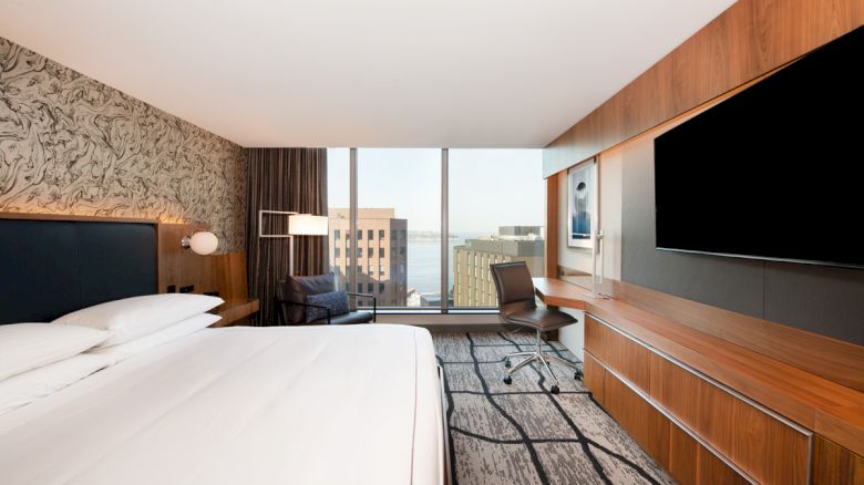 A modern hotel room with a large bed, city view through windows, a desk with a chair, mounted TV, and contemporary furnishings.