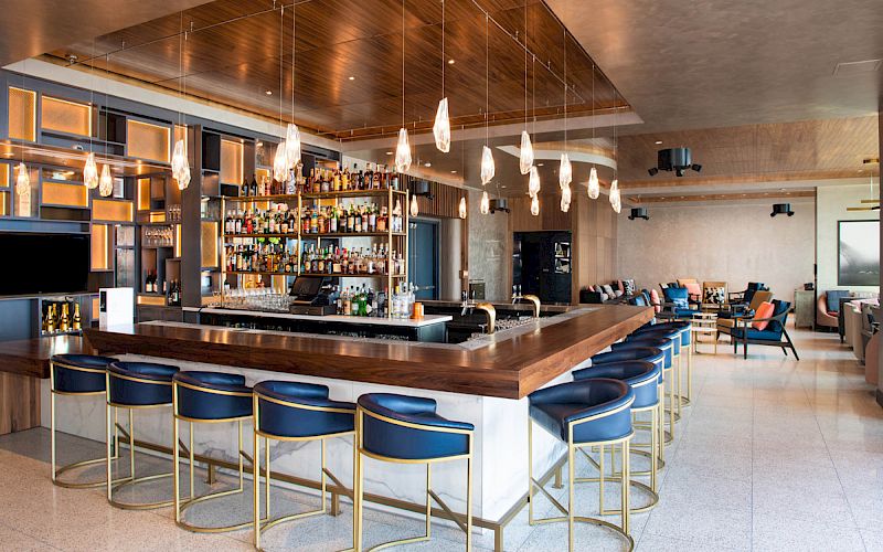 A sleek, modern bar setup with blue stools, hanging lights, and a fully stocked bar in an upscale, spacious lounge area with additional seating.