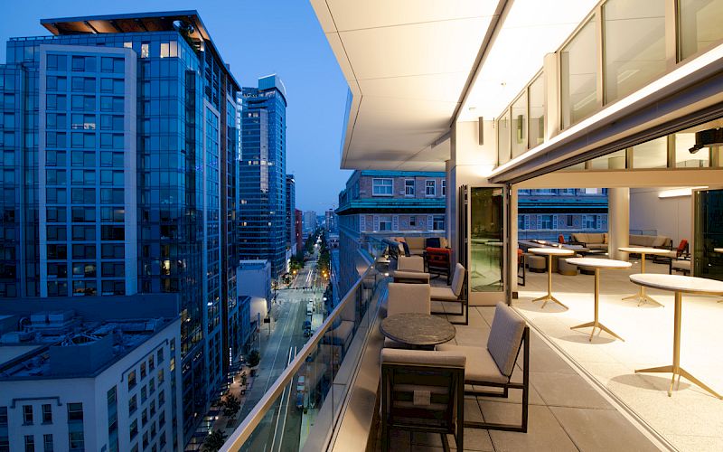 A modern urban balcony at dusk with seating and tables, overlooking a city street lined with high-rise buildings and lights.