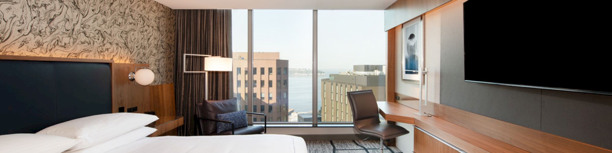 A modern hotel room with a large bed, wall-mounted TV, desk, chair, armchair, window with city view, and stylish decor.